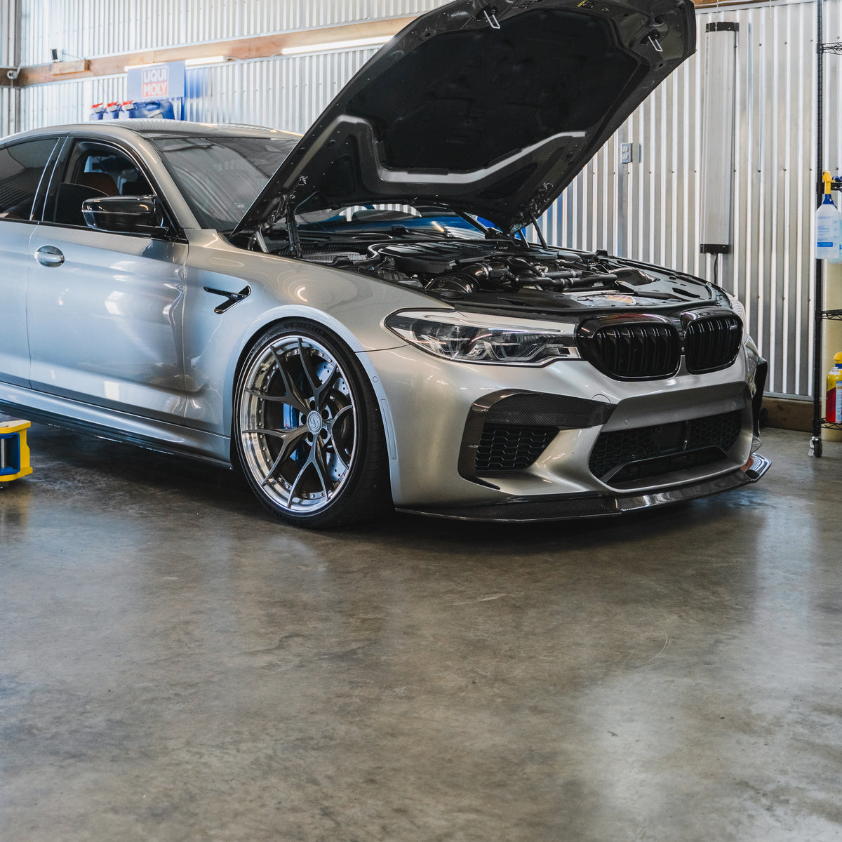 Silver BMW M5 with its hood open in a workshop bay.